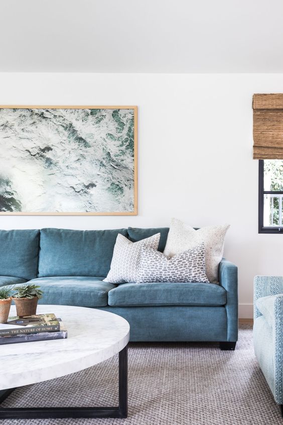 a lovely modern coastal living room with a blue sofa, a light blue chair, a marble top table, a coastal artwork and a neutral rug