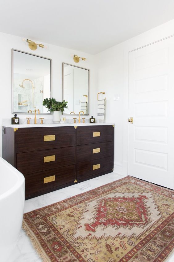 a modern farmhouse bathroom with a double dark-stained vanity with gold fixtures and gold handles, mirrors and gold sconces over them