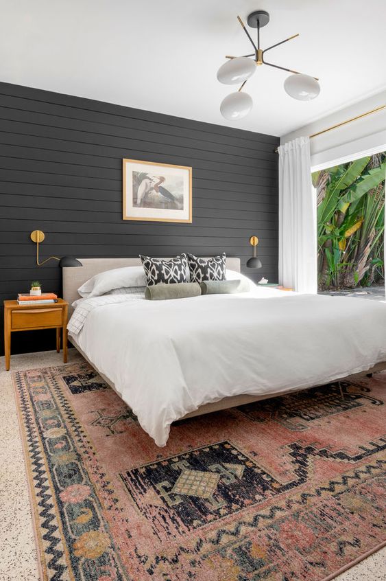 a modern farmhouse bedroom with a black shiplap accent wall, a neutral bed, a printed rug, light-stained nightstands and a cool chandelier