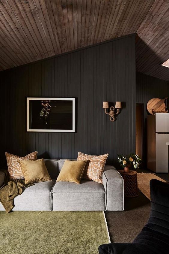 a moody living room with a soot shiplap wall, a grey low sofa, a yellow rug, an artwork and some bright pillows