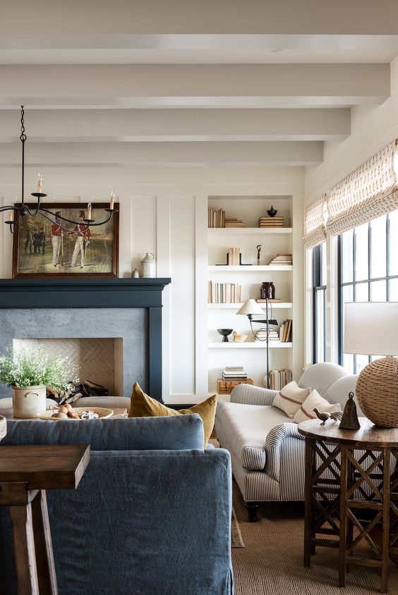 a neutral farmhouse living room with wooden beams, built-in shelves, a fireplace with a black mantel, a white sofa, a blue one, a coffee table