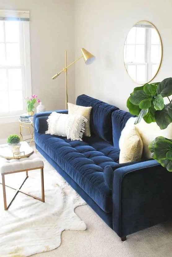 a neutral living room with a navy sofa with neutral pillows, a round mirror in a gold frame, a creamy upholstered bench, a gold floor lamp and a side table