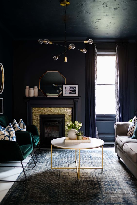 a soot living room with a fireplace clad with gold and soot panels, a neutral sofa, a coffee table, green chairs, a stylish chandelier