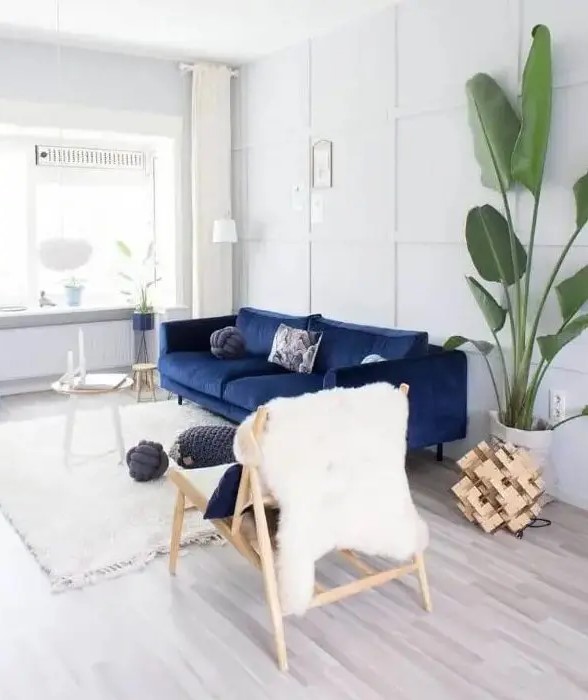 a pretty light-filled living room done in white and creamy, with a navy sofa, a couple of side tables, a wooden chair, a potted tree and some wood