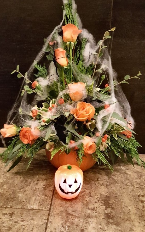 a pumpkin vase with greenery and bright orange blooms covered with spiderwebs is a chic floral arrangement for Halloween