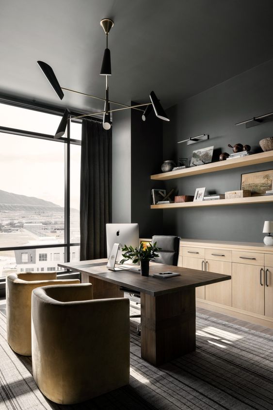 a refined home office with soot walls, a glazed wall, a dark-stained desk, neutral and a black chair, a black chandelier and light-stained cabinets
