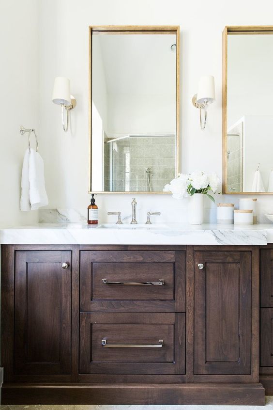 a refined neutral bathroom with a dark-stained vanity, a white stone countertop, mirrors in gilded frames and neutral sconces