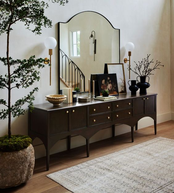 a refined vintage dark-stained console table with artwork, books and vases, a large mirror and a potted tree next to it