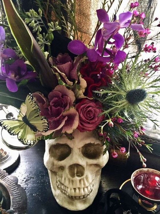 a skull centerpiece with a lush floral arrangement in purple, pink, green, with thistles and butterflies plus large leaves