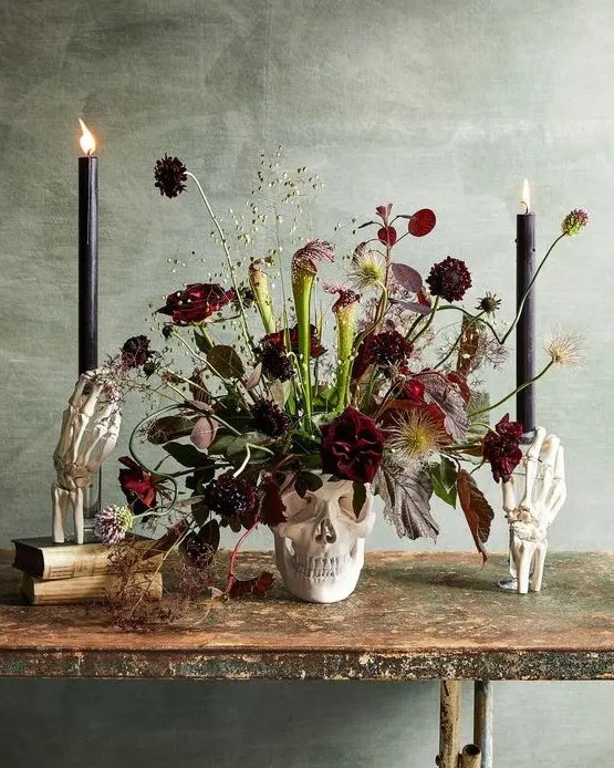 a skull vase with burgundy blooms, dark and usual leaves, greenery and black candles for Halloween