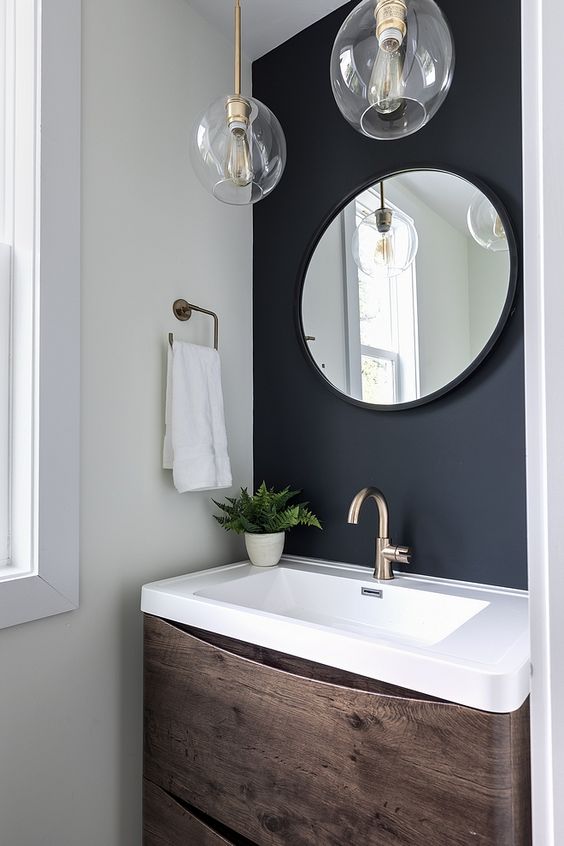 a small powder room with a soot accent wall, a timber vanity with a stone sink, a round mirror and pendant lamps