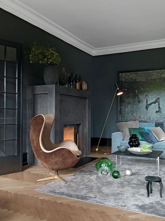 a soot living room with a firpelace, a dark artwork, a blue sofa and colorful pillows, a brown egg-shaped chair and some vases