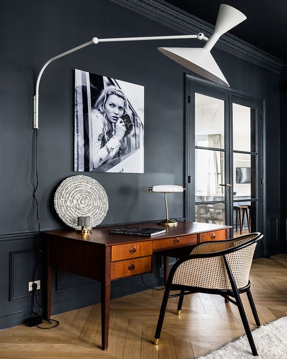 a soot space with a vintage rich-stained desk and a cane chair, a large wall lamp and an artwork