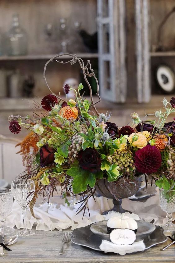 a sophisticated Halloween flower arrangement of orange, burgundy and deep purple blooms, some greenery, herbs and twigs