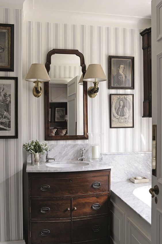 a vintage bathroom done with striped wallpaper, a dark-stained vanity, a gallerywall and a mirror in a stained frame, a couple of vintage sconces