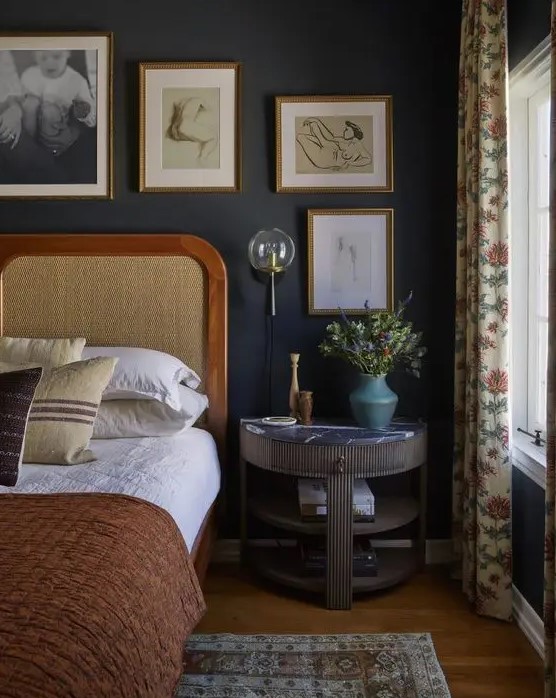a vintage moody bedroom with soot walls, a bed with a cane headboard, a fluted nightstand, a gallery wall and printed textiles