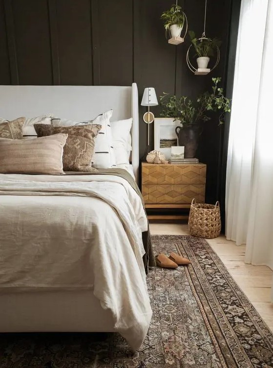 a welcoming moody bedroom with a soot accent wall, a neutral upholstered bed with neutral bedding, a printed rug, pendant plants and an inlay nightstand