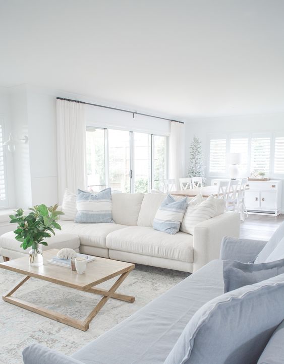 an airy coastal living room with a white and pale blue sofa, printed pillows, a wooden folding table, greenery and a neutral printed rug