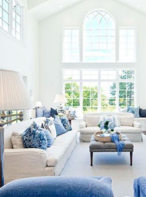 an airy living room with a double-height ceiling, neutral sofas, blue pillows, poufs and an ottoman, blue blooms
