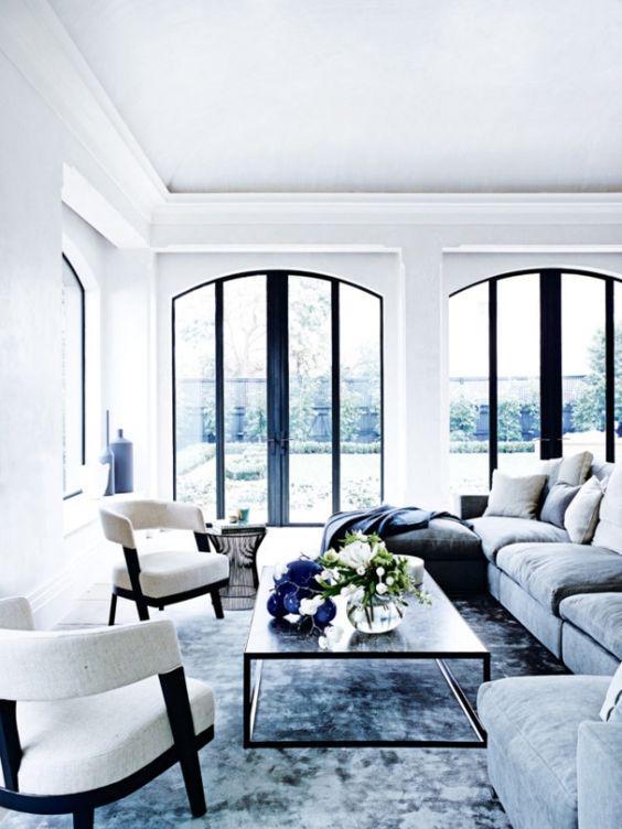 an airy living room with arched windows with black framing, a blue rug, grey blue sofas and white chairs, a coffee table