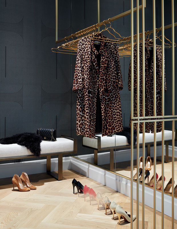 an elegant closet with a soot patterned wall, a mirror wall, a neutral bench, gold railings and clothes hangers