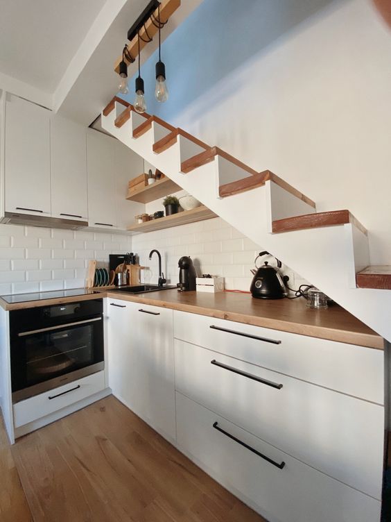 a Scandinavian kitchen with sleek cabinets, butcherblock countertops, a white subway tile backsplash and a staircase over the kitchen