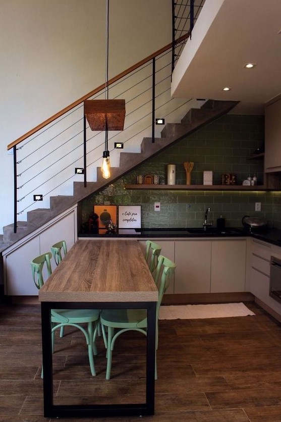 a catchy modern kitchen placed under the staircase, with a green tile backsplash, greige lower cabinets, black countertops and an open shelf