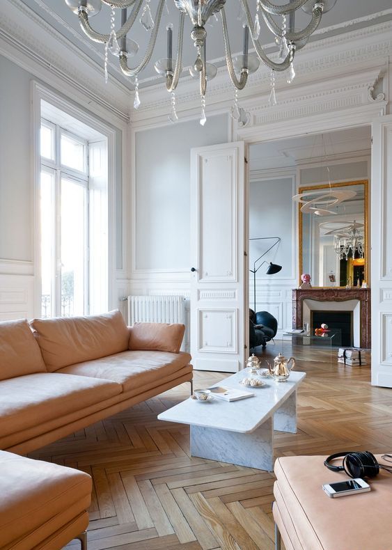 a beautiful Parisian chic living room with grey walls and herringbone floors, a peachy sofa and ottoman, a marble table, a fireplace and a black chair
