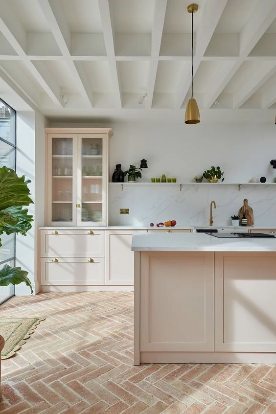 a beautiful modern peachy kitchen with terracotta herringbone tiles on the floor, a white stone backsplash and countertops and gold pendant lamps