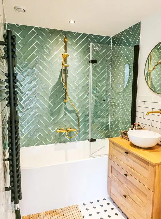 a cozy bathroom with green herringbone and white skinny tiles, a stained vanity, an oval tub and gold touches
