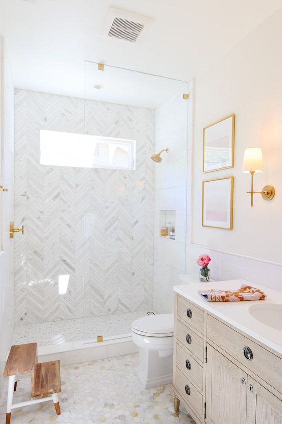 a cozy neutral bathroom with marble herringbone tiles, a stained vanity, a stool, some lamps and artwork