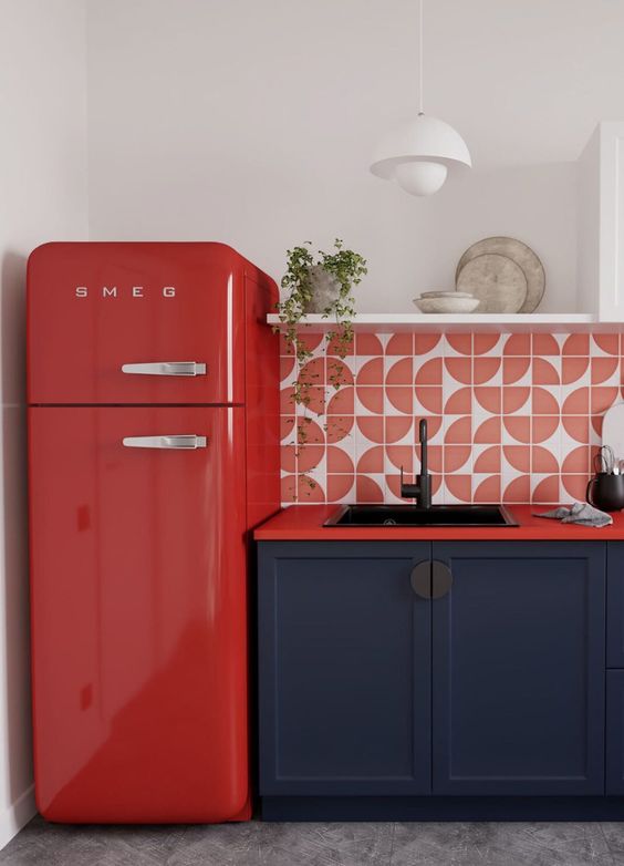 a lovely navy kitchen with red countertops, a red printed tile backsplash and a red fridge plus an open shelf and some greenery