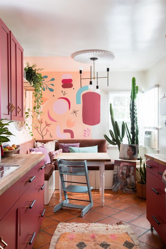 a matte red kitchen with butcherblock countertops, bright wall art, a banquette seating and a table plus some potted plants