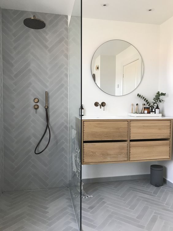 a modern bathroom done with grey herringbone tiles, a stained vanity, a round mirror and dark fixtures