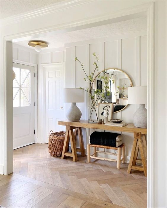 a neutral modern farmhouse space with a light-stained herringbone floor, a trestle console table, a stool and a large mirror, some lamps