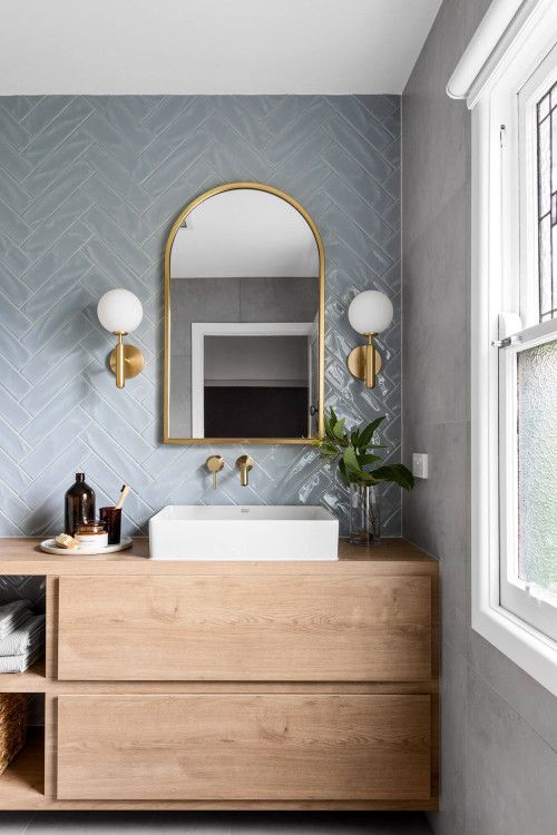 a pale blue herringbone tile wall, a stained vanity, a mirror with lamps and a white sink for a modern bathroom