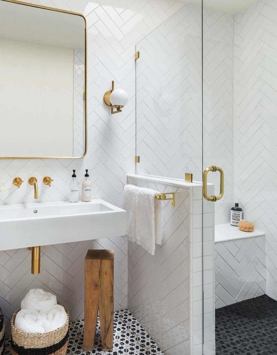 a small bathroom clad with white herringbone tiles, a shower space with a bench, a wall-mounted sink and gold fixtures