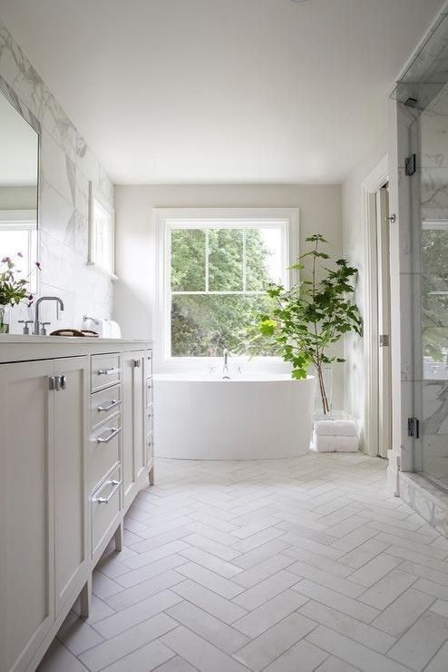 a sophisticated white bathroom with white marble on the walls and a white herringbone tile floor, a large vanity and a large tub