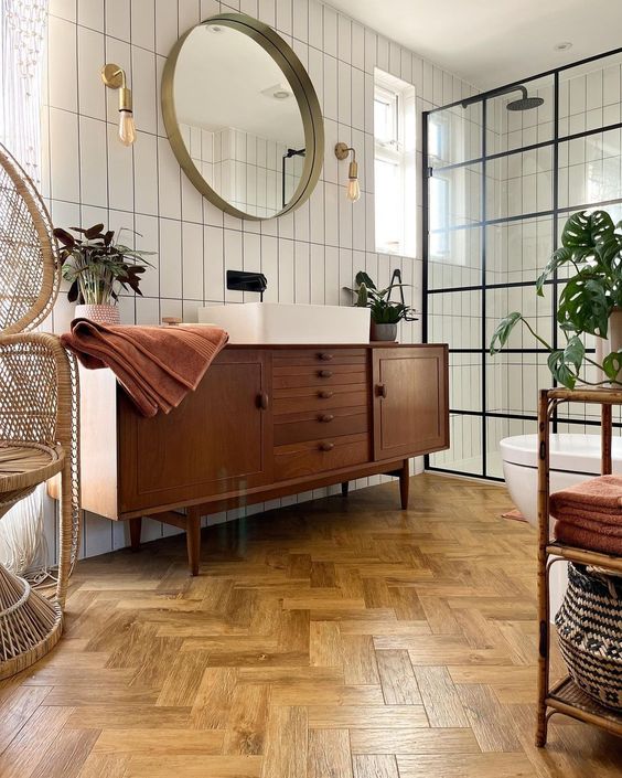 a stylish mid-century modern bathroom with white skinny tiles and a herringbone floor, a stained vanity, a papasan chair and a shower