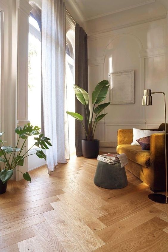 a stylish off-white living room with a herringbone floor, a mustard chair, a green pouf, potted plants and a gold floor lamp