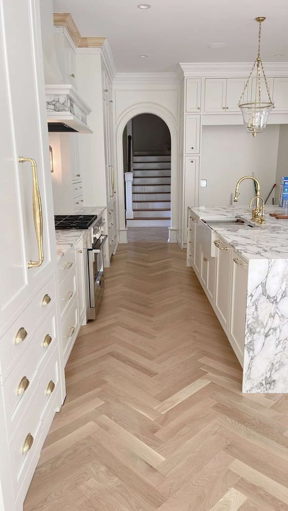 an exquisite white kitchen with shaker style cabinets, a large kitchen island with a white stone countertop and a light-stained herringbone floor