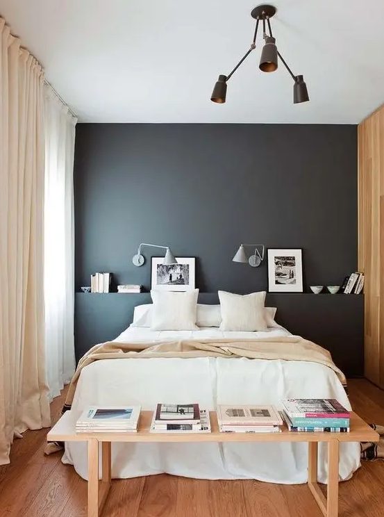 a chic modern bedroom with a black accent wall, a bed with neutral bedding, a bench with books and a chandelier
