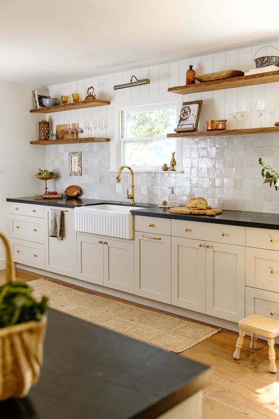 a cozy farmhouse kitchen with tan cabinets, black countertops, a pearly Zellige tile backsplash, stained shelves and gold fixturesHome