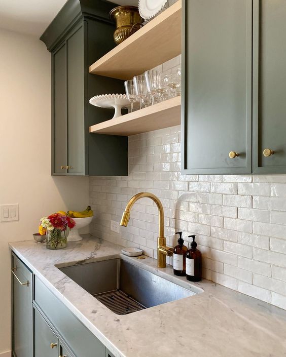 a green kitchen with neutral stone countertops, a white Zellige tile backsplash and gold fixtures