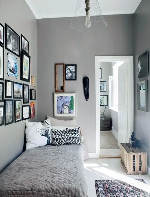 a grey narrow bedroom with a small bed, a crate for storage and gallery walls and a pendant lamp