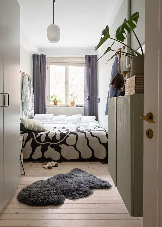 a narrow bedroom with a bed and printed bedding, mauve curtains, a rug, a green cabinet and potted plants and books