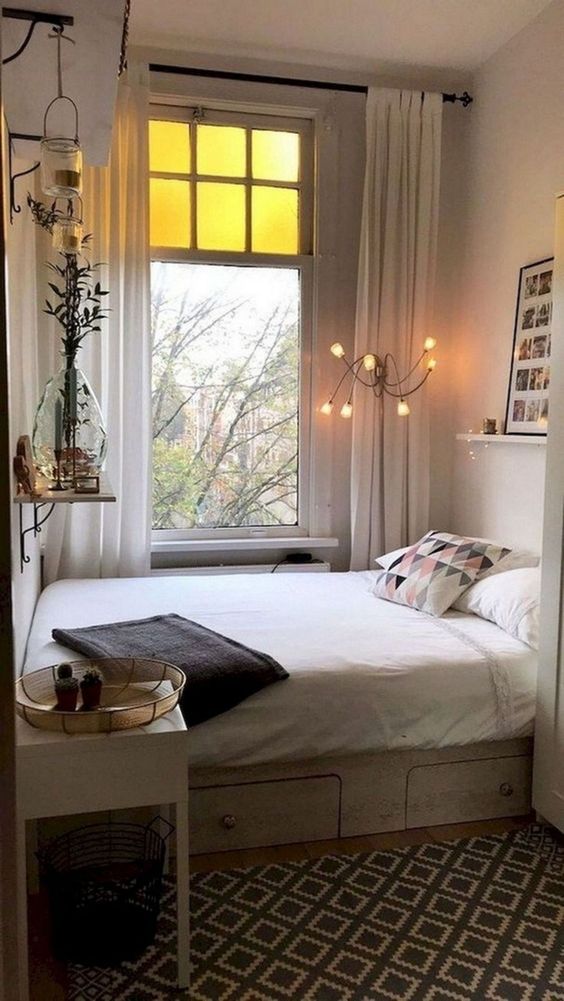 a narrow bedroom with a bed placed horizontally, neutral bedding, a nightstand, some shelves and pretty decor
