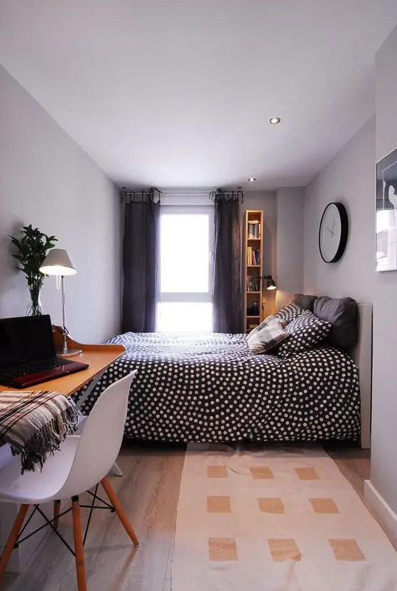 a narrow grey bedroom with a bed with printed bedding, a narrow and tall bookshelf, grey drapes, a printed rug, a wall-mounted desk and a white chair