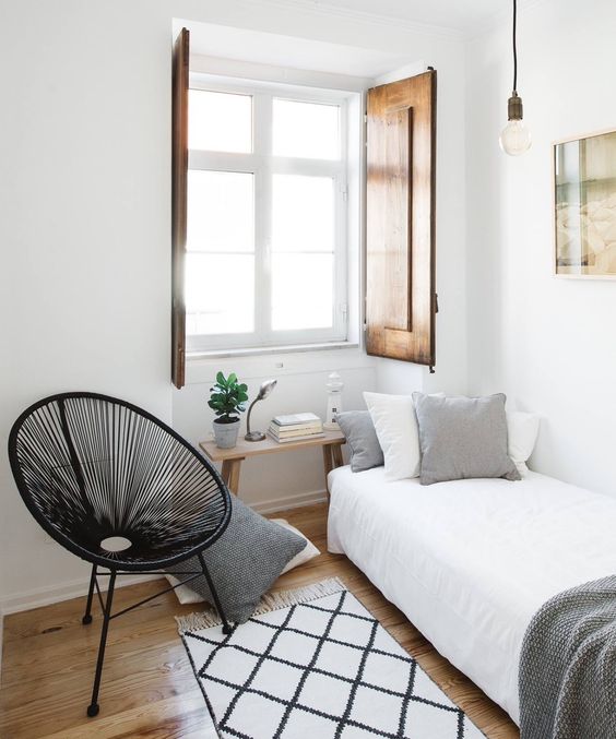 a narrow guest bedroom with a bed and neutral bedding, a round chair, a bench as a nightstand and wooden shutters
