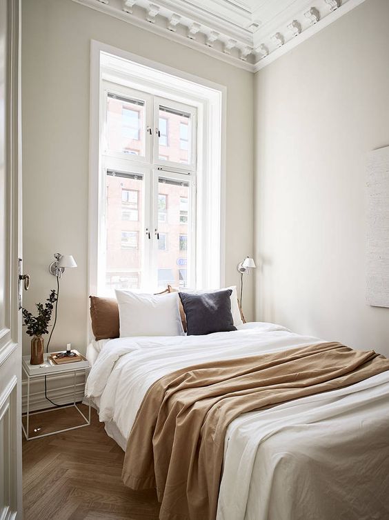 a narrow sleeping space with a bed and neutral and colored bedding, nightstands and wall lamps